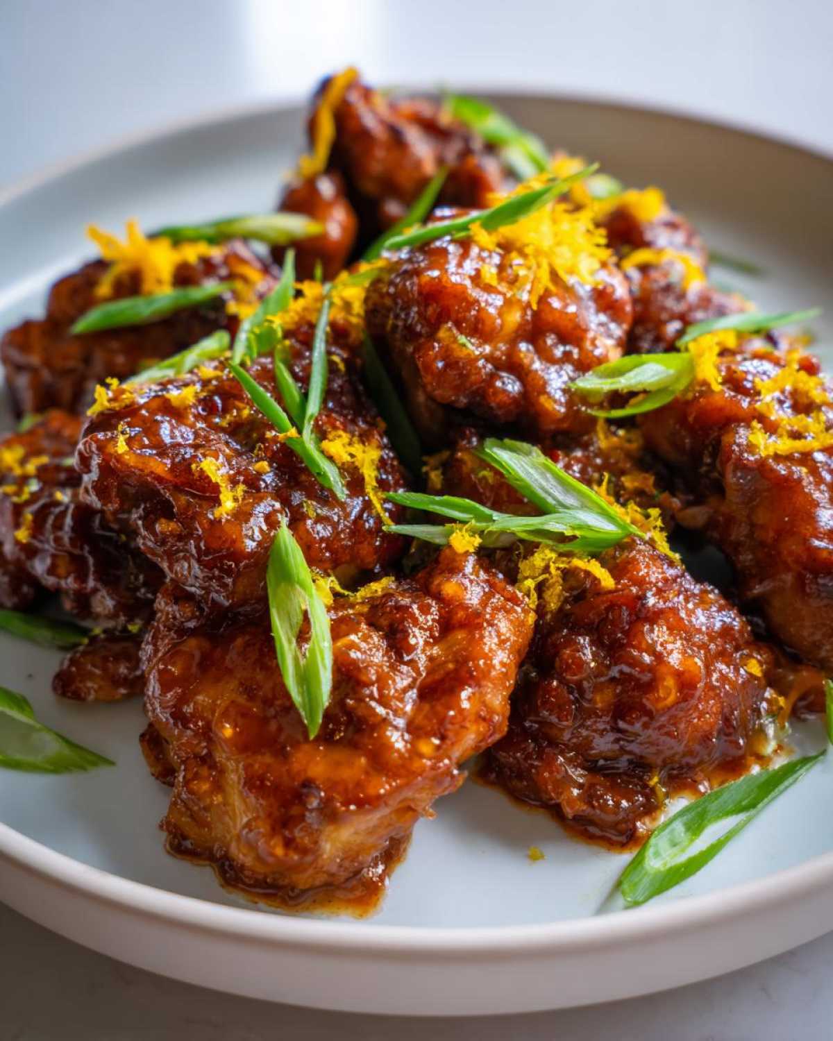 Close-up of Sticky Orange Chicken Thighs coated in a glossy sauce, garnished with bright orange zest and sliced green onions.