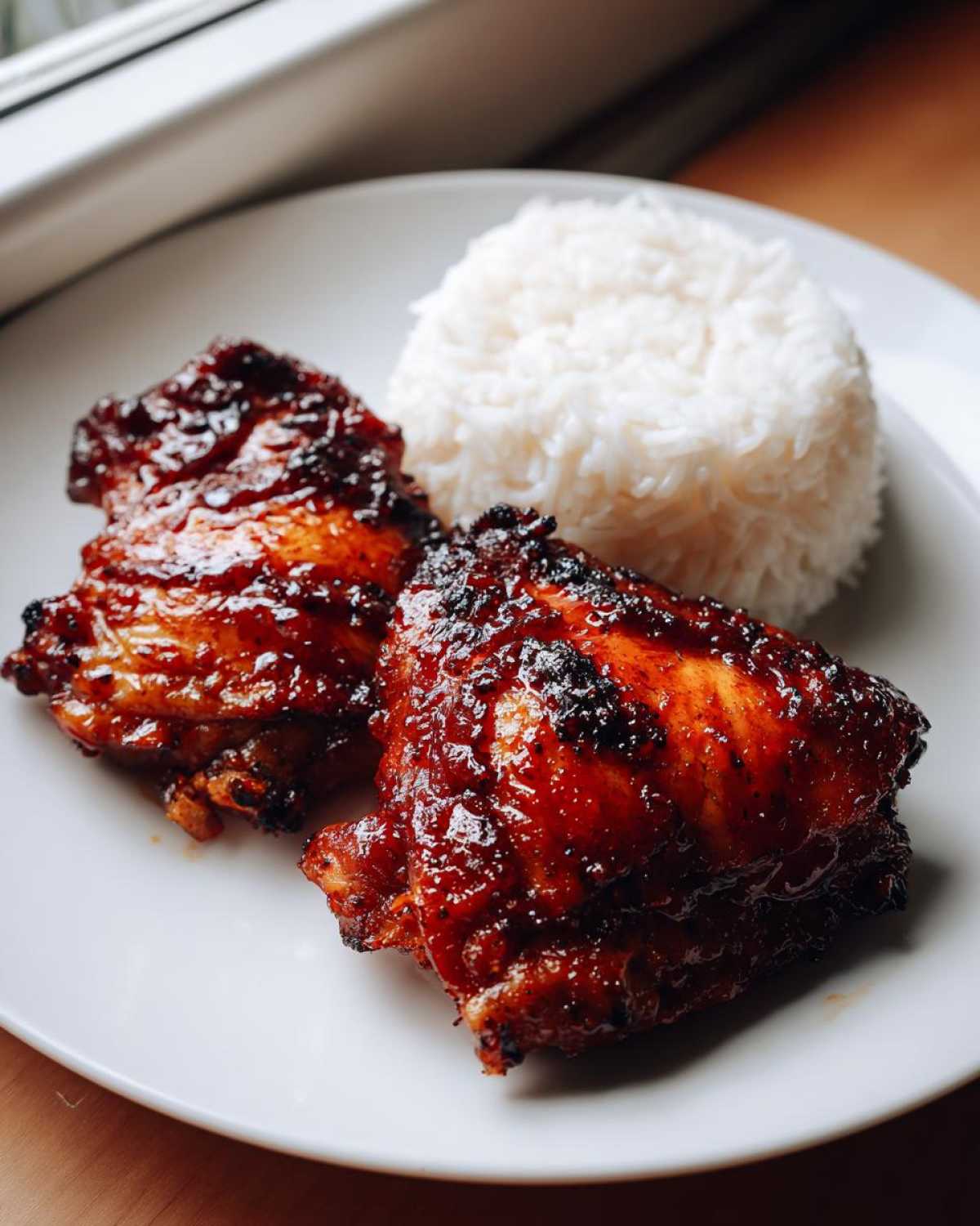 Two glazed Sticky Sweet Chili Chicken Thighs served next to a mound of white rice on a white plate.