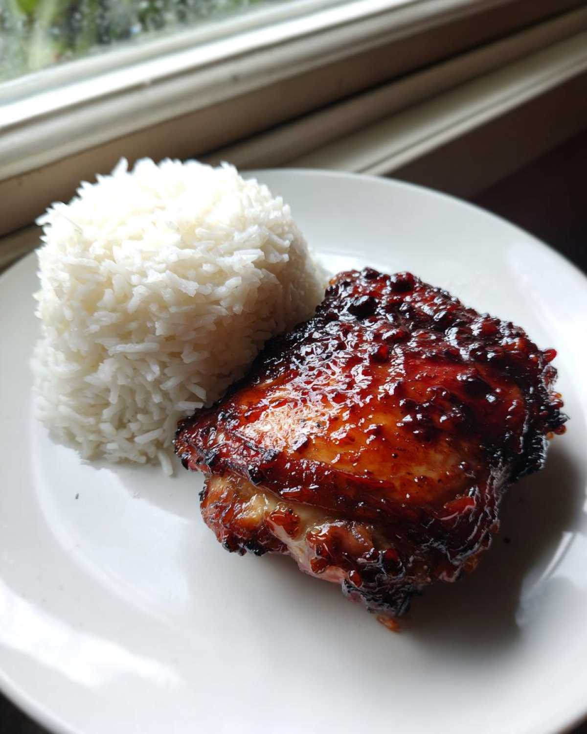 A perfectly cooked Sticky Sweet Chili Chicken Thighs served next to a mound of fluffy white rice on a white plate.