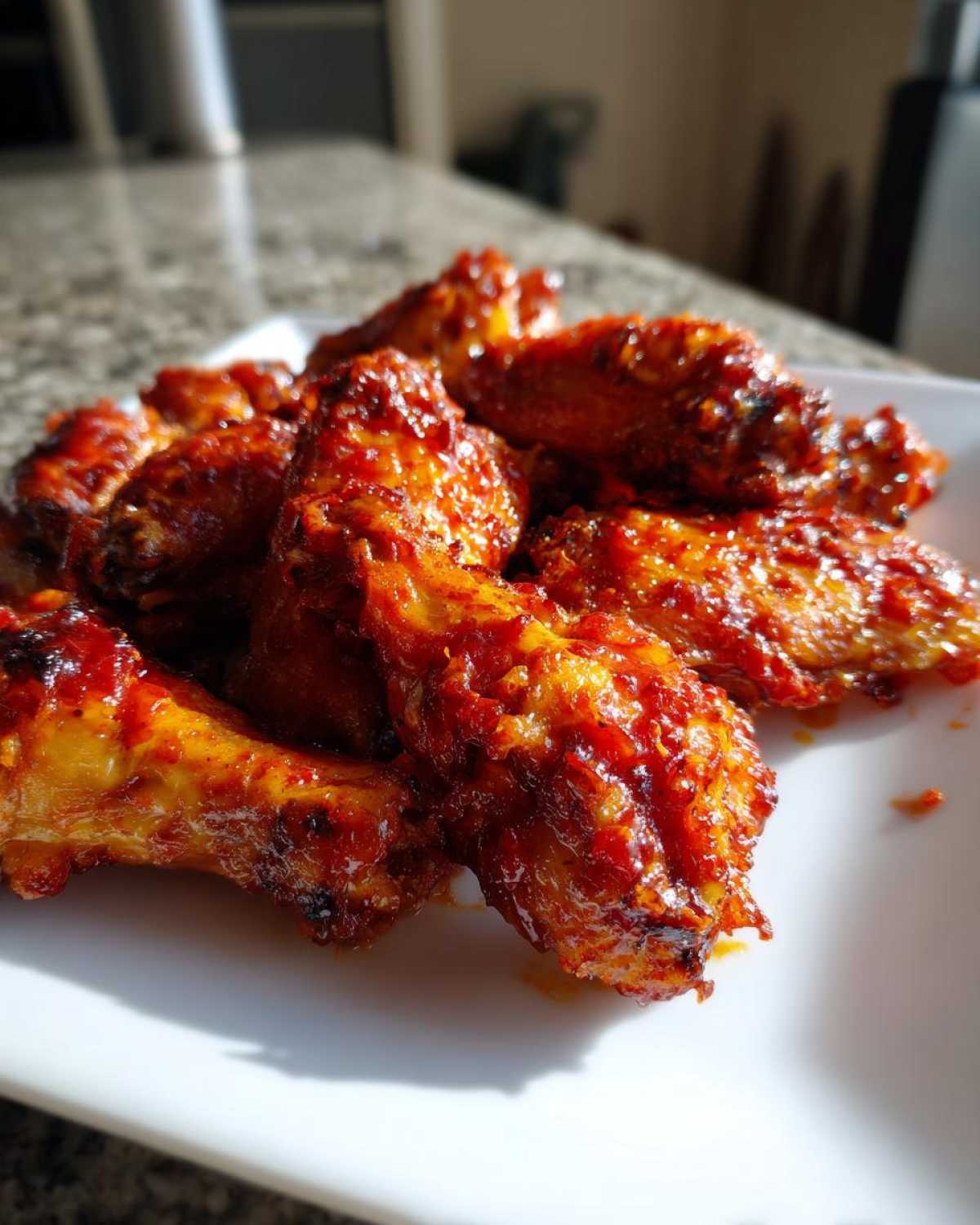 A close-up of several oven-baked Sticky Sweet Chili Chicken Wings coated in a glossy, deep red glaze on a white plate.