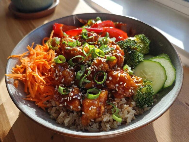 A close-up of a Sweet Chili Chicken Bowl featuring glazed chicken pieces over brown rice, topped with sesame seeds, green onions, and crunchy vegetables.